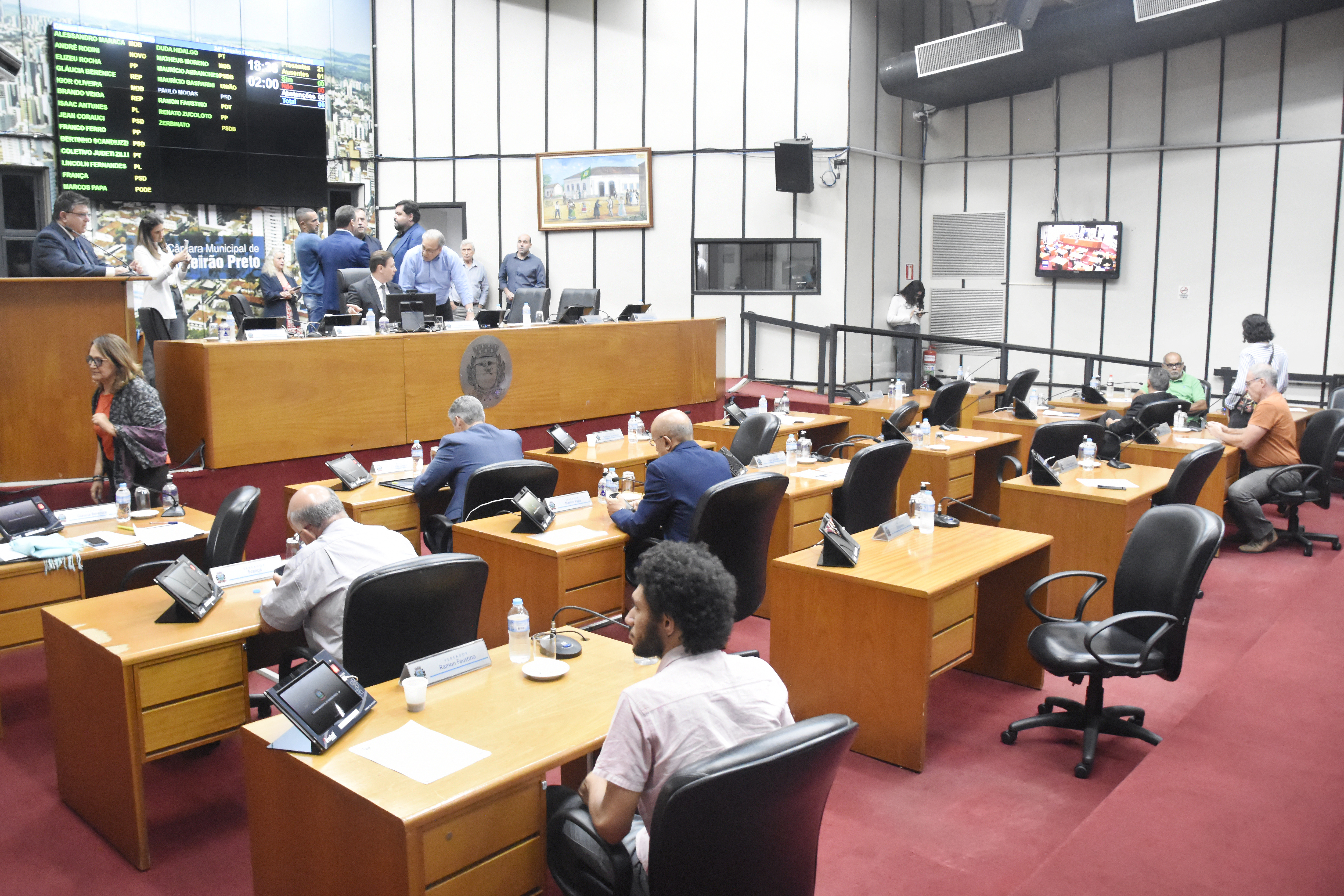 Imagem aberta do plenário de frente pra Mesa diretora. Alguns vereadores de costas.