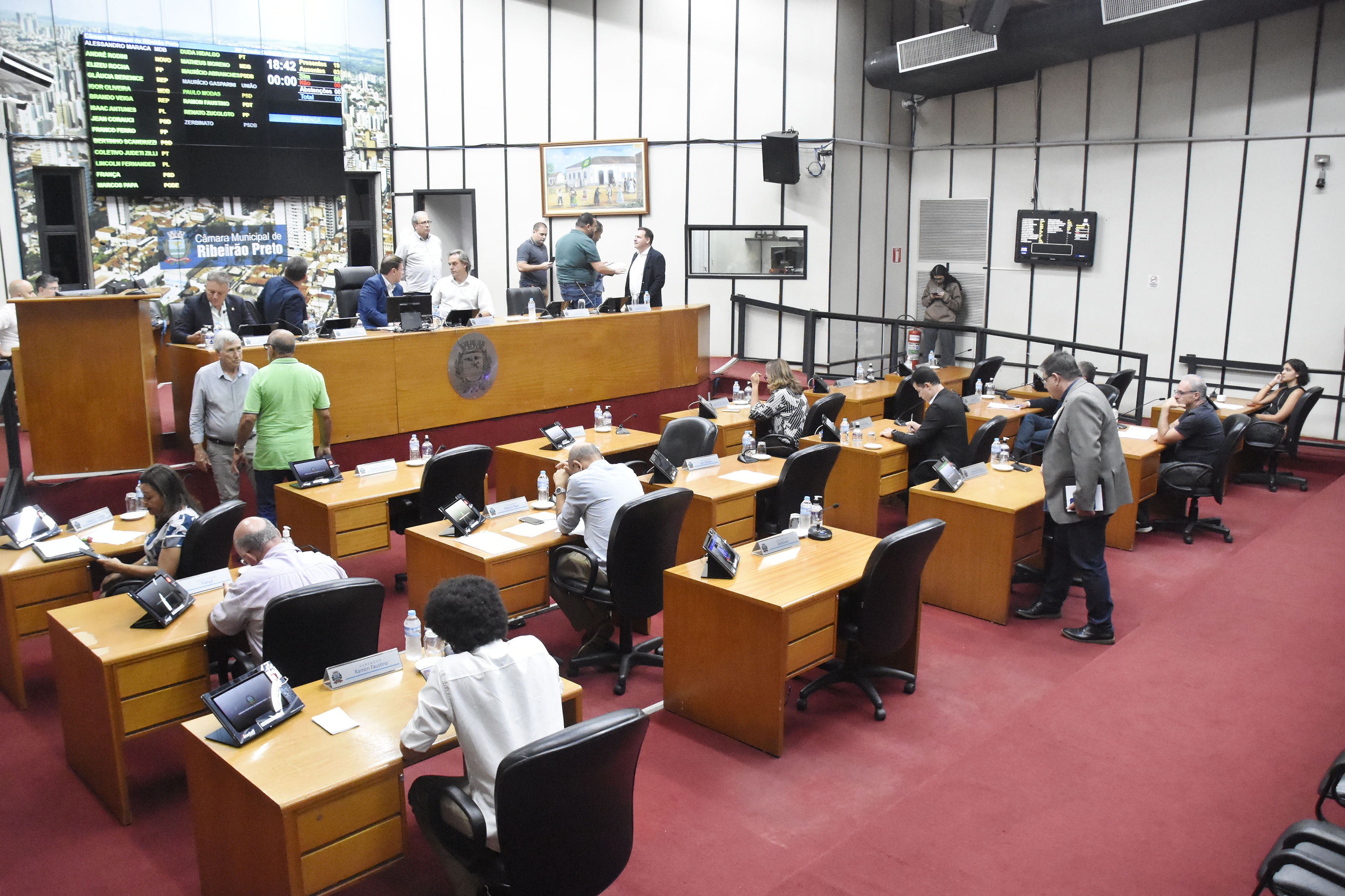 Imagem aberta do Plenário da Câmara Municipal, mostrando a mesa diretora de frente e outros vereadores de costas