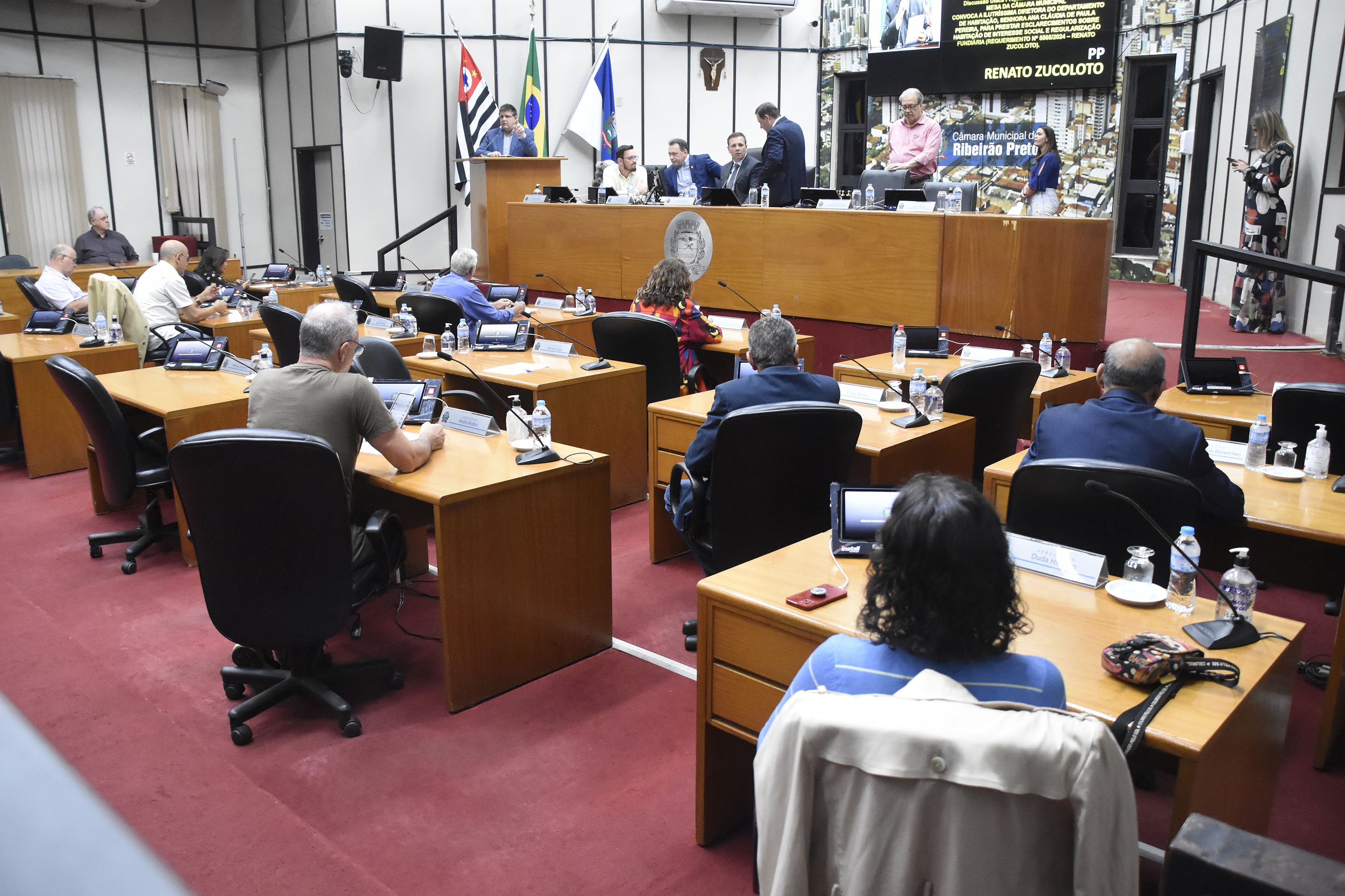 Imagem aberta do Plenário com a mesa diretora de frente e demais vereadores de costas